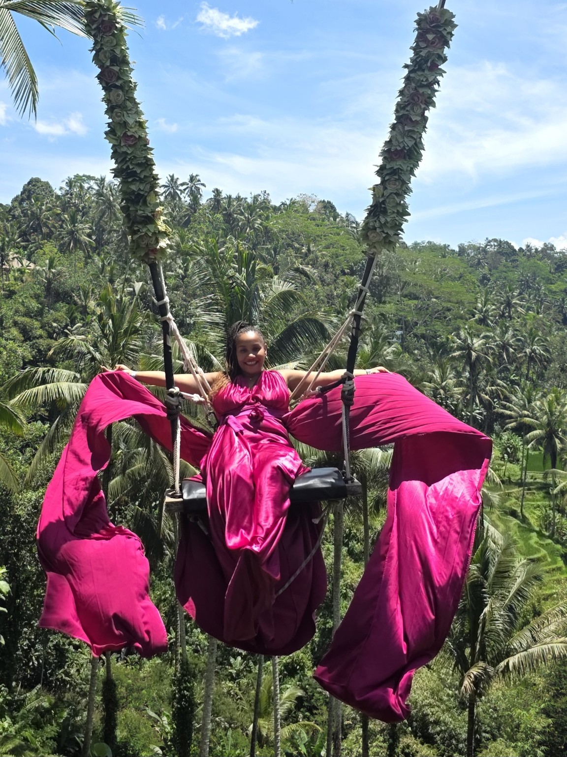 Leslie in a flowing pink dress on a tropical swing above a lush landscape